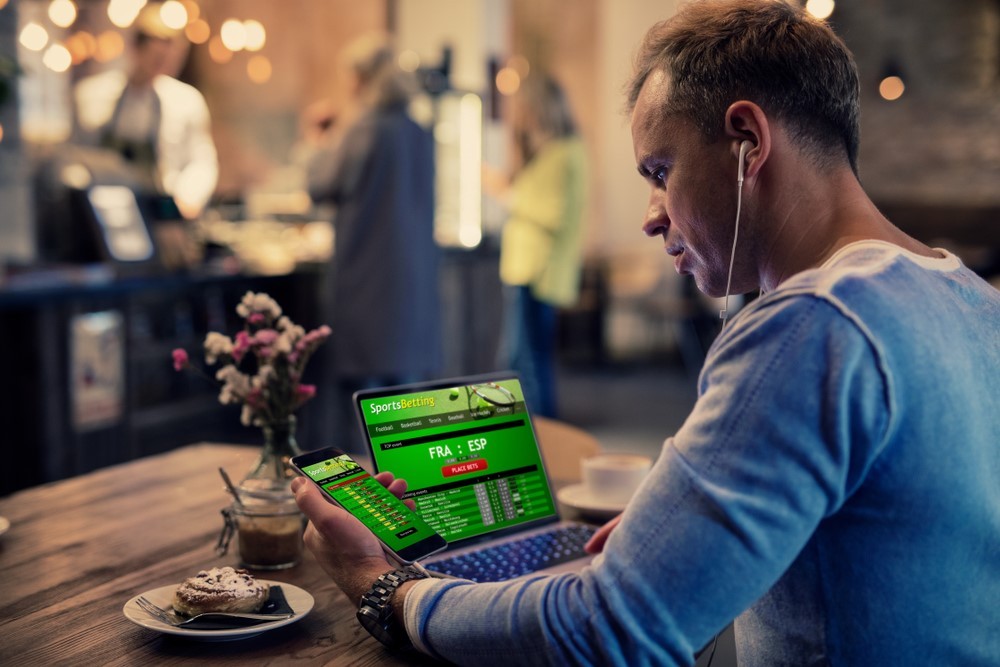 Ein Mann verfolgt verschiedene Sportwetten auf einem Laptop und Smartphone in einem Café